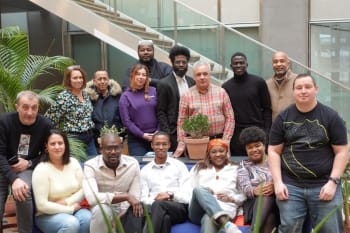 A group of fifteen people pose indoors on a blue couch and nearby chairs in front of a staircase, with potted plants around them.