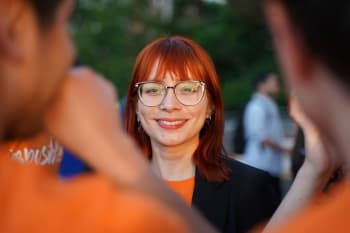 A person with red hair and glasses smiles at the camera, surrounded by people wearing orange shirts, outdoors.