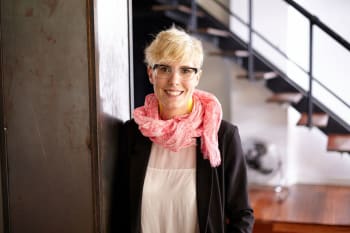 A person with short blonde hair, glasses, and a pink scarf smiles while standing indoors near a staircase.