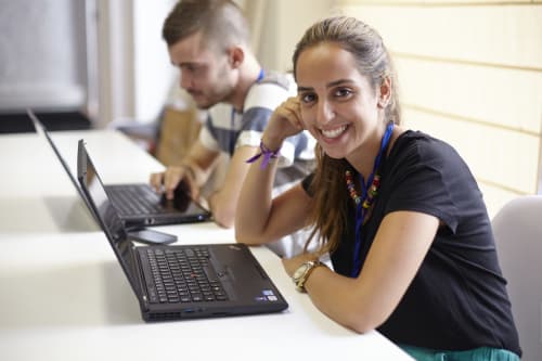 Two people sit at a table using laptops. The woman in the foreground is smiling at the camera, while the man in the background is focused on his work.