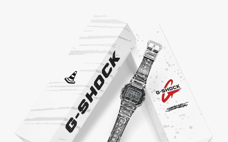 Black and white illustrated G-Shock digital watch with patterned strap next to white G-Shock branded boxes.
