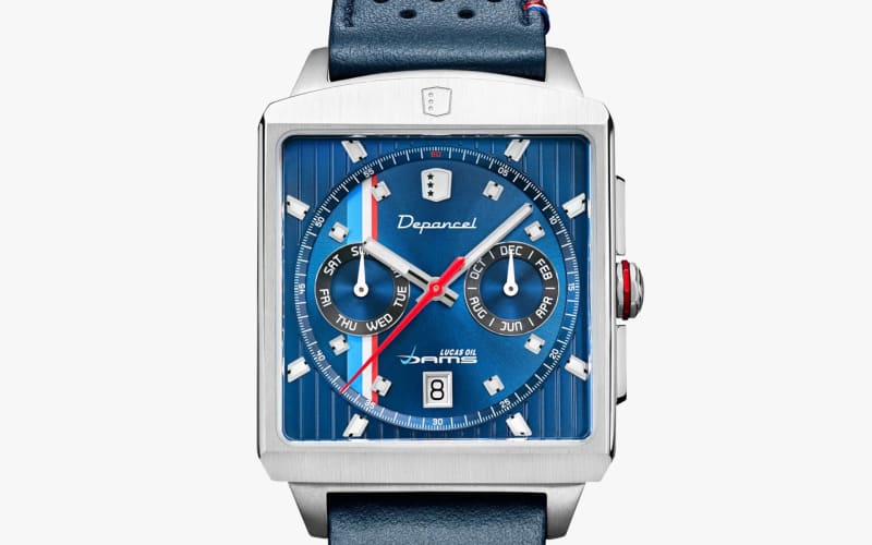 Square silver watch with blue perforated leather strap and blue dial featuring day, month subdials, and red second hand.