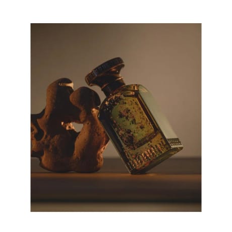 Flacon de parfum par Anfas (Ghala Extrait) avec un bouchon doré orné et du verre vert, 100 ml, à côté d'une sculpture en pierre rustique.