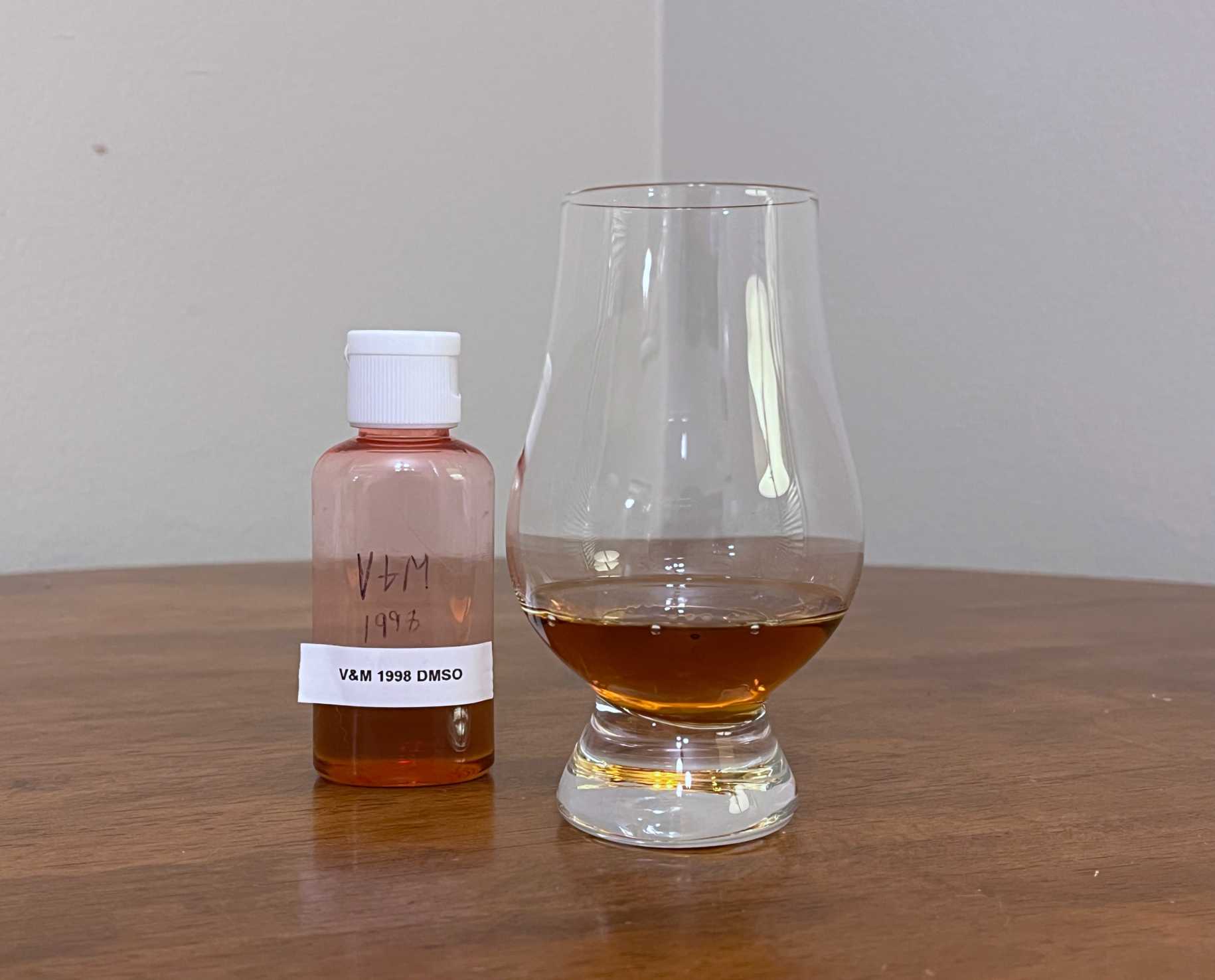 A sample bottle of Valinch & Mallet Bellevue 1998 next to a glass of rum on a table