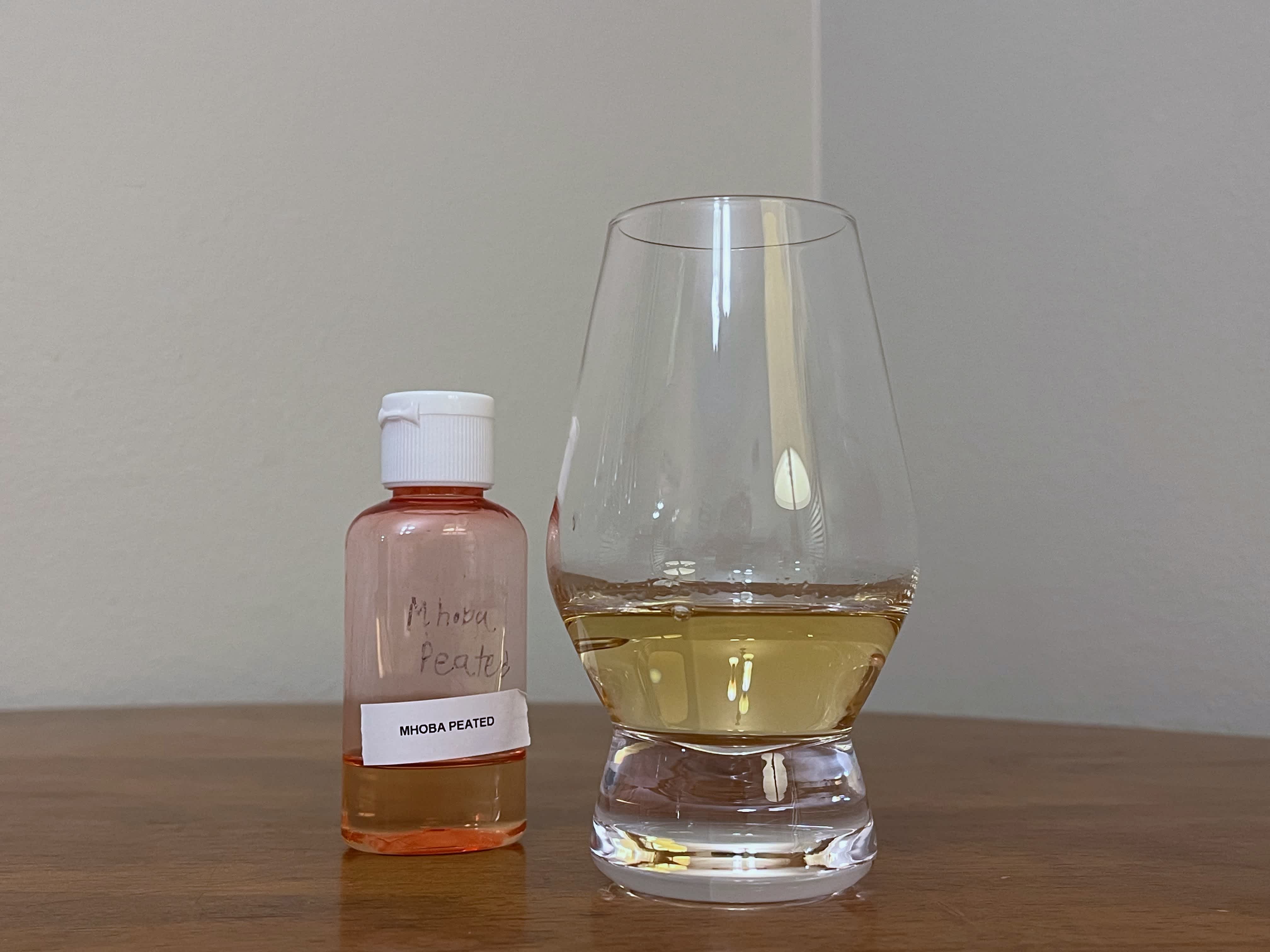 Sample bottle of Mhoba Peated on a tabletop next to a glencairn with rum poured in it