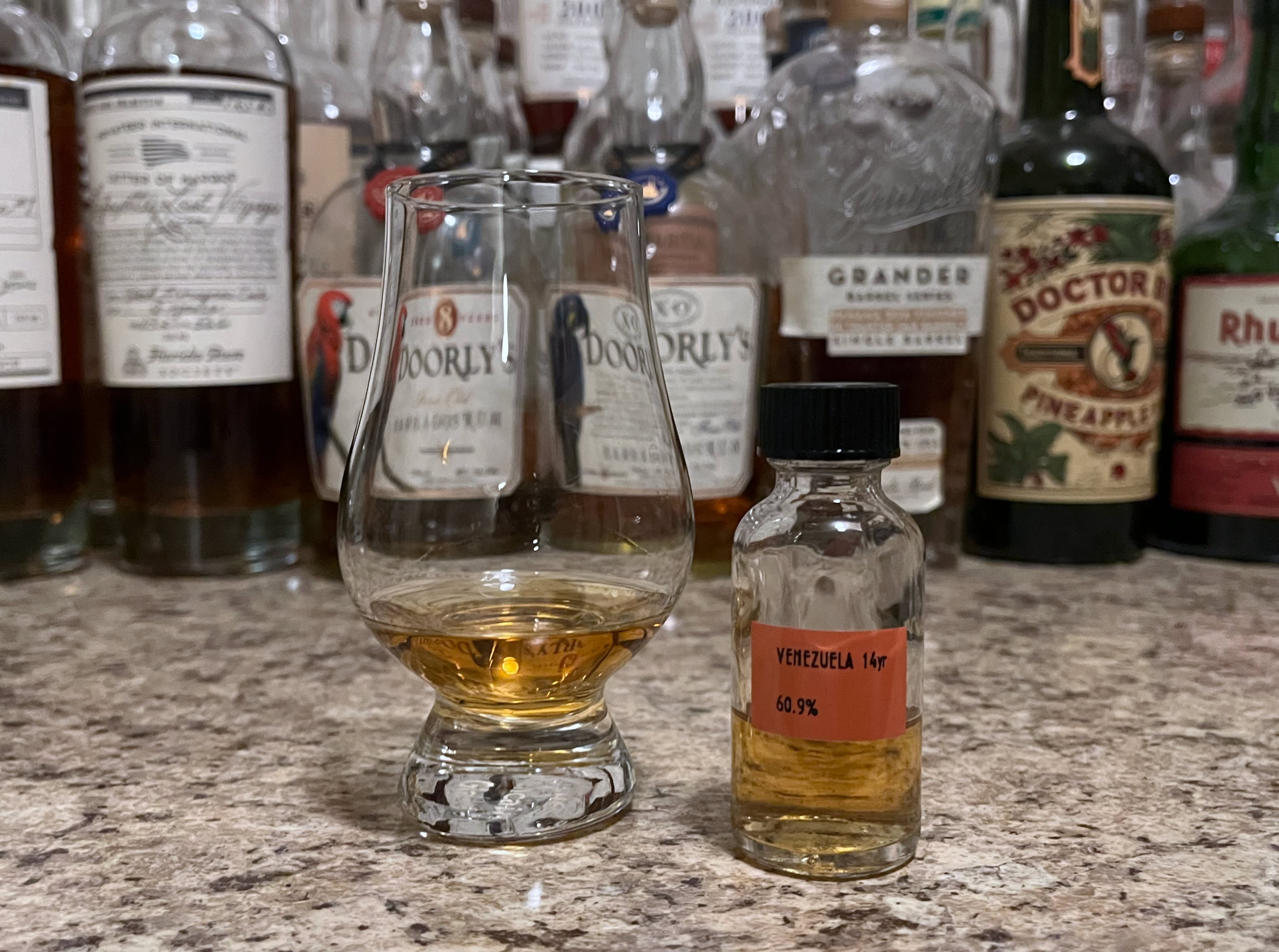 A sample bottle of Raising Glasses Venezuela next to a glencairn of rum on a kitchen counter