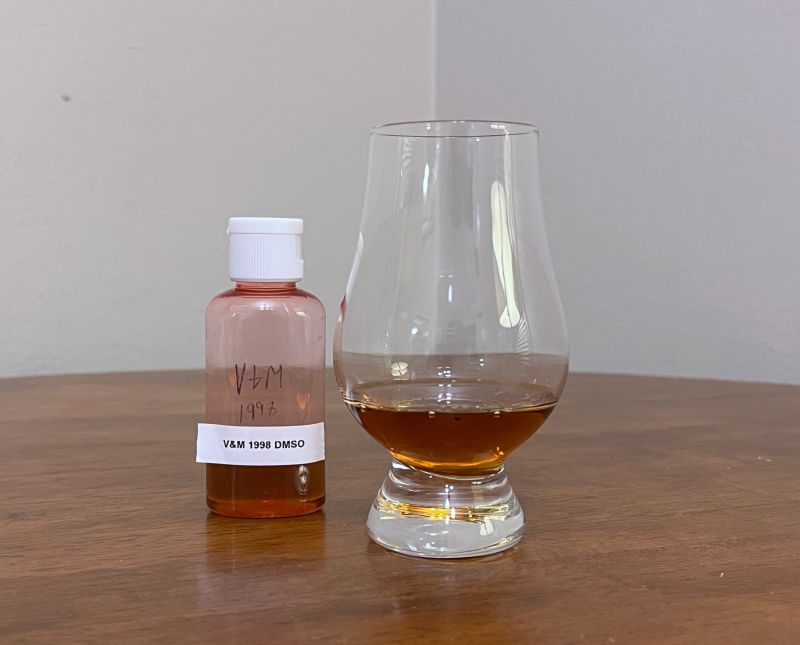 A sample bottle of Valinch & Mallet Bellevue 1998 next to a glass of rum on a table