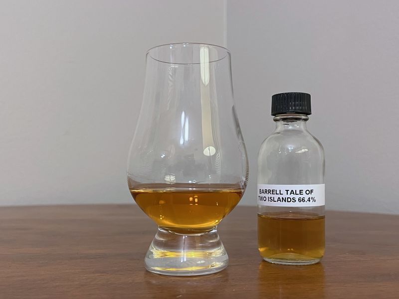 A sample bottle of Tale of Two Islands next to a glass of rum on a tabletop