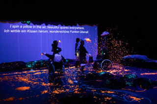 Under UV light, cushions, clothing, a blue curtain and orange confetti glow. Six performers dance and twirl confetti. Roy Fischer as Crispy spins around in his wheelchair. The projected lyrics appear on the curtain: ‘I spin a pillow in the air, beautiful sparks everywhere.’