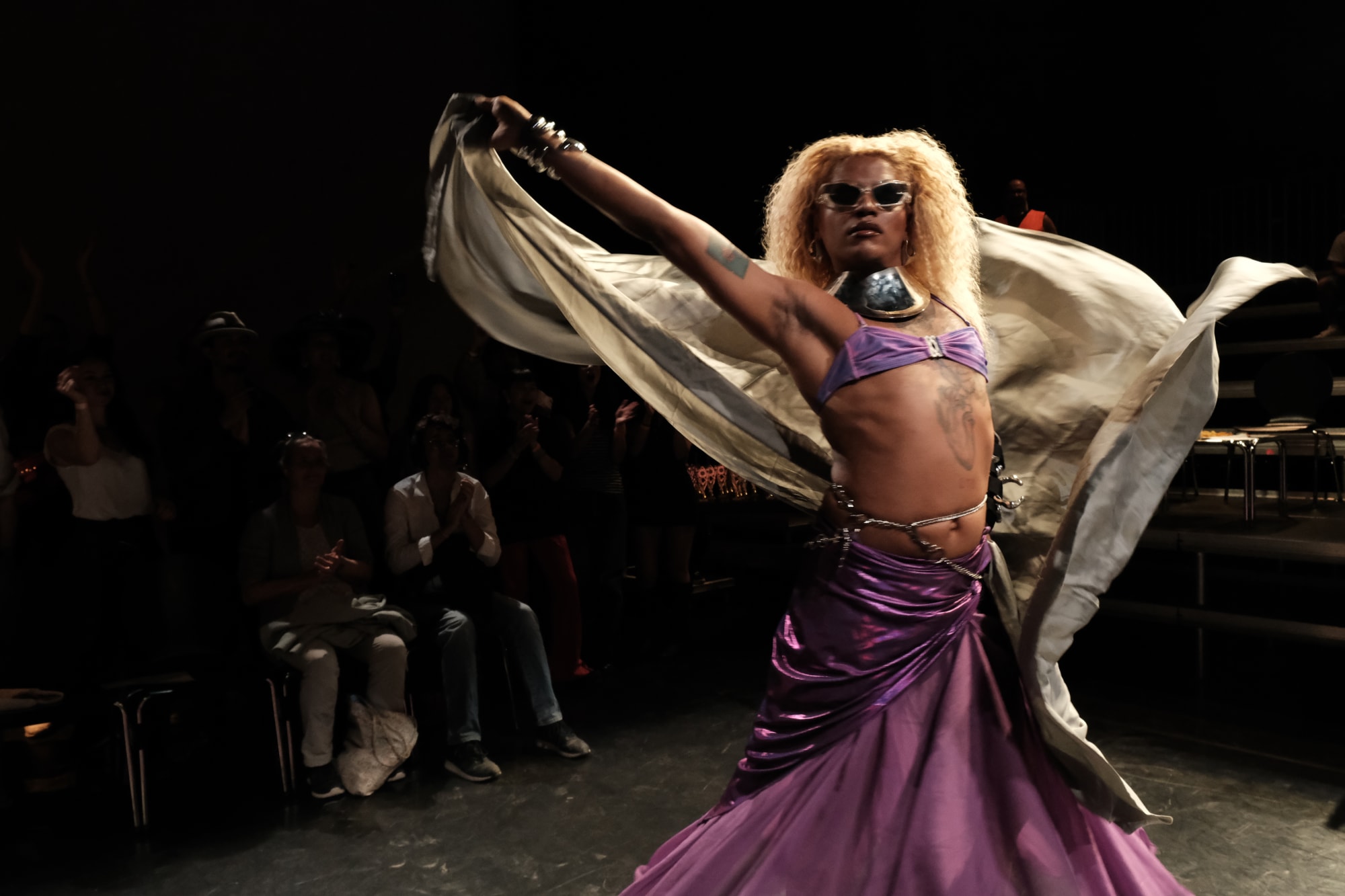 Person bei einer Performance, trägt Sonnenbrille, schwingt einen Schal, trägt violetten Rock und Top.