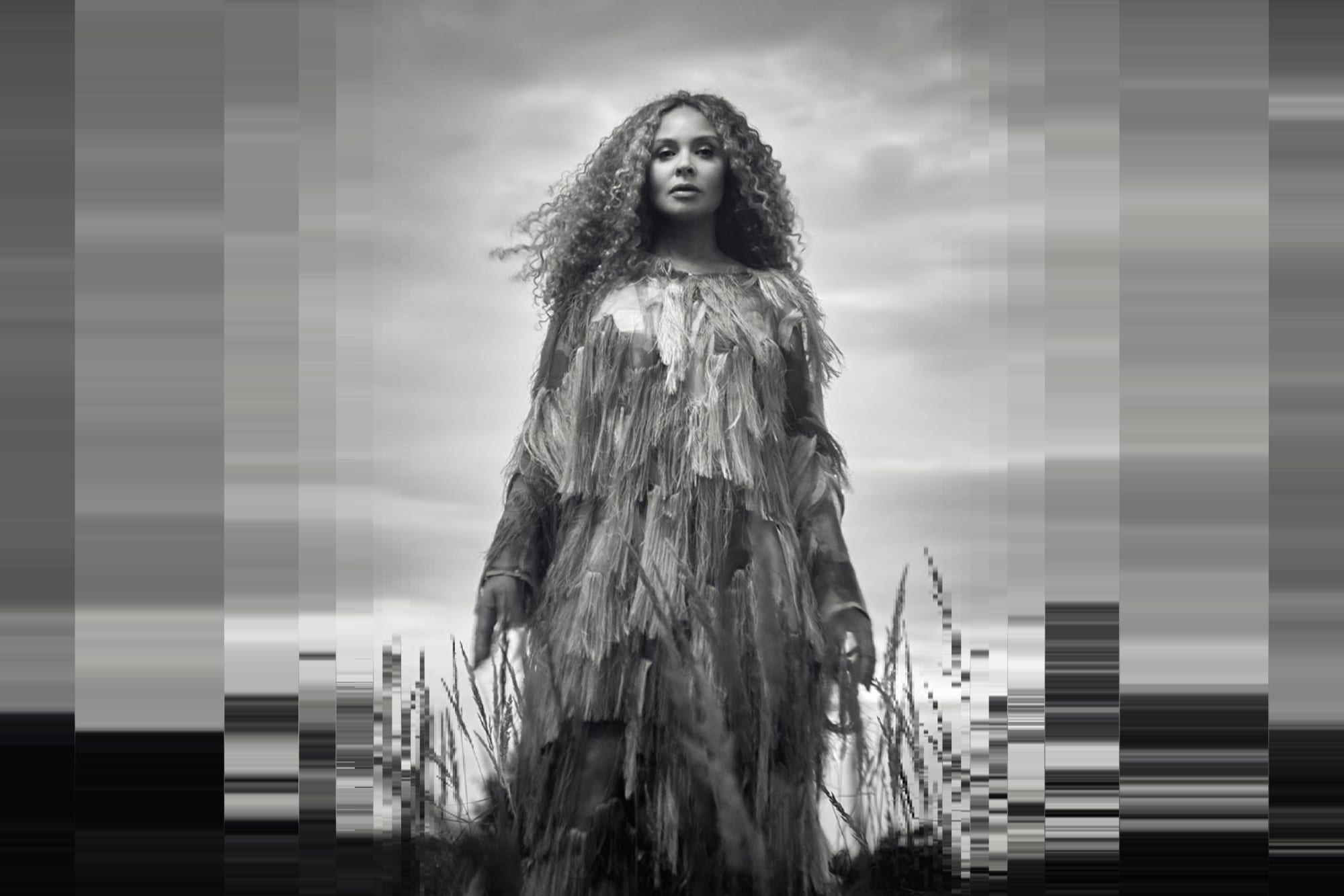 Joy Denalane stands in front of a cloudy sky in the middle of the picture, which is held in shades of gray. Her long hair is moved by the wind. The image dissolves into vertical pixels at the edges.