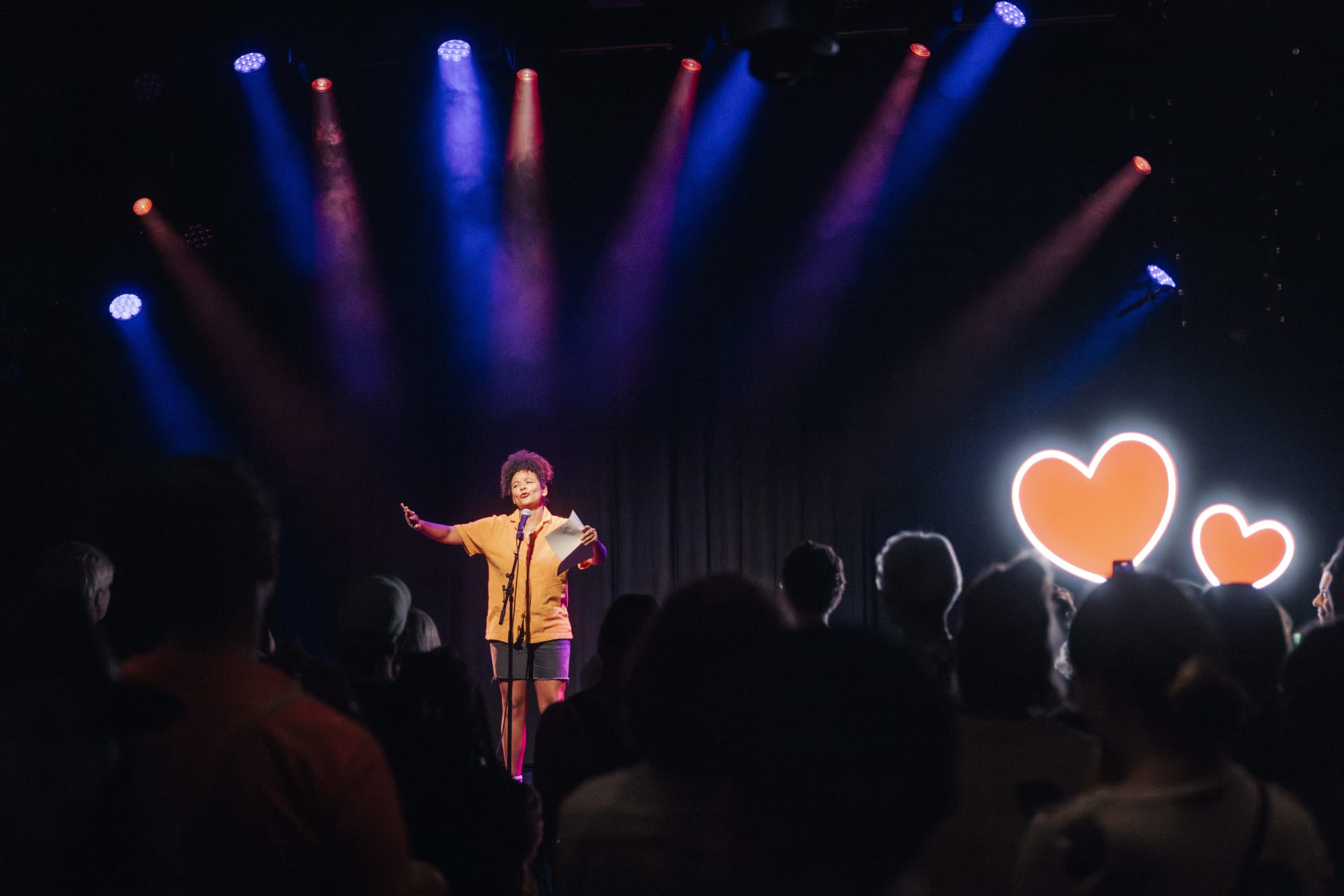 Fatima Moumouni holding papers on stage under colorful lights, addressing a crowd. Large glowing hearts are displayed in the background, creating a warm ambiance.