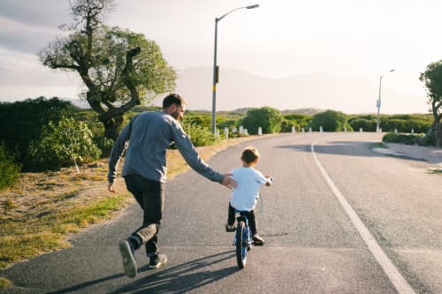 Dad teaching son to ride bike on asphalt road surrounded by trees