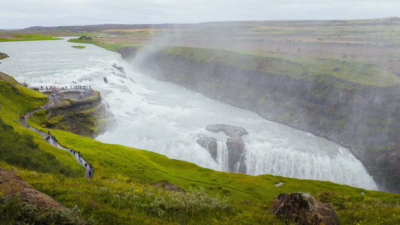 Gullfoss waterfall from the top of a overlook point surrounded by mist from the waterfall next to lush green grass and a few tourists up close to the waterfall