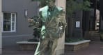 Thumbnail about Statue of a balding businessman, left hand in pant's pocket, right hand pointing horizontally.