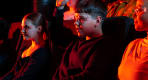 Thumbnail about A young boy wearing safety glasses raises his hand while sitting in a dark theater alongside other children and adults, illuminated by the r