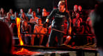Thumbnail about A female presenter in a "Lady Lava" shirt pours glowing orange molten lava with a metal rod in front of an engaged audience watching in awe.