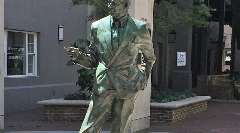 Statue of a balding businessman, left hand in pant's pocket, right hand pointing horizontally.