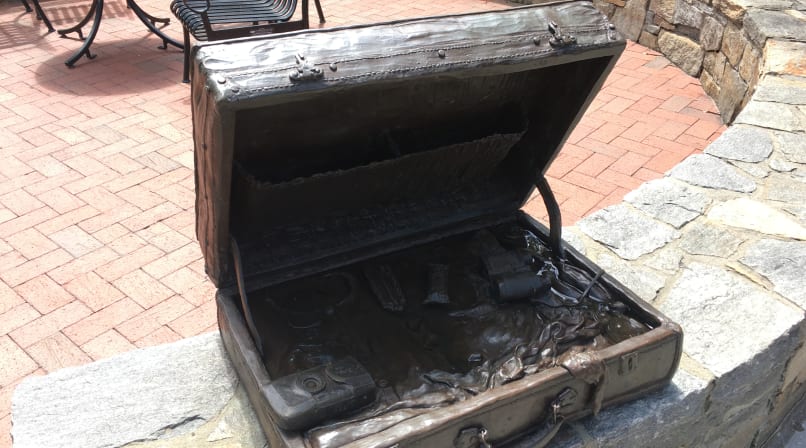 Sculpture of an old, open suitcase containing a camera, binoculars, sunglasses, snacks, and clothes. It sits on a ledge.