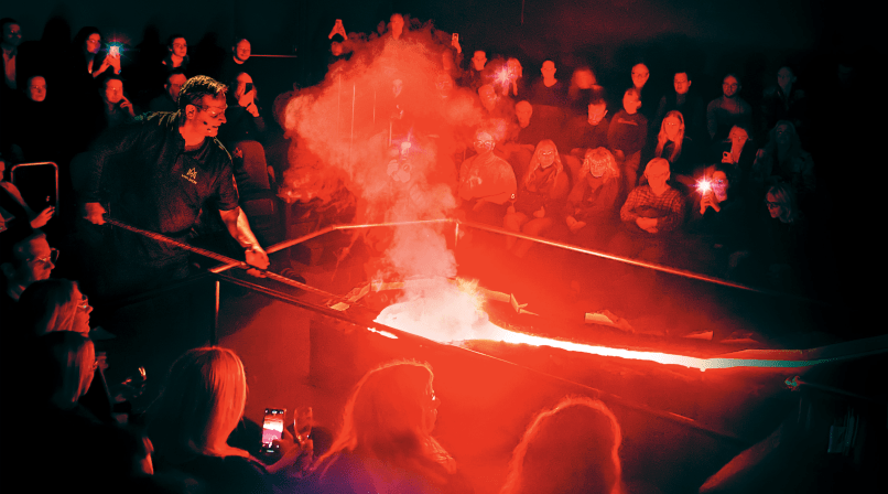 A Lava Show presenter uses a tool to manipulate a stream of glowing molten lava while a full audience watches under red light and rising ste