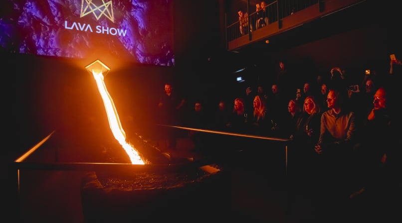 Molten lava flows down a steel channel in a dark showroom with the "Lava Show" logo projected on a screen, while the audience watches from b