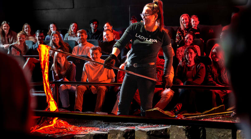 A female presenter in a "Lady Lava" shirt pours glowing orange molten lava with a metal rod in front of an engaged audience watching in awe.