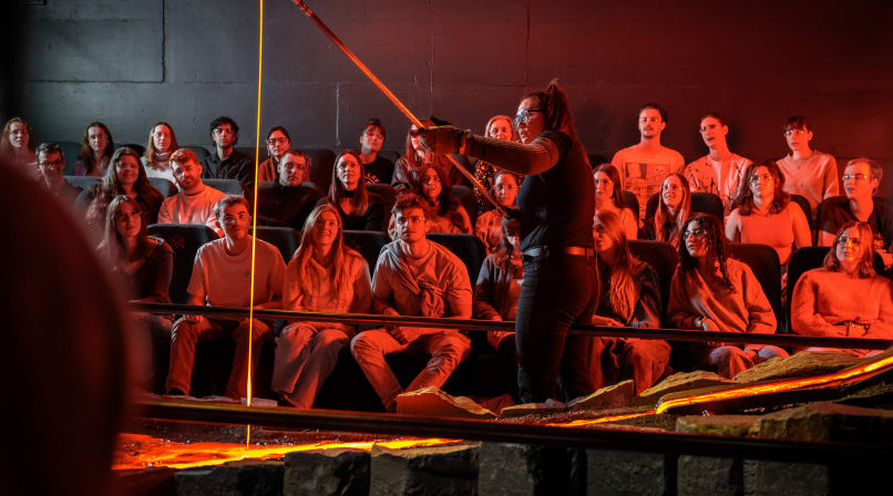 A close-up of a lava stream pouring from a tilted metal container under intense orange lighting with a person standing nearby.