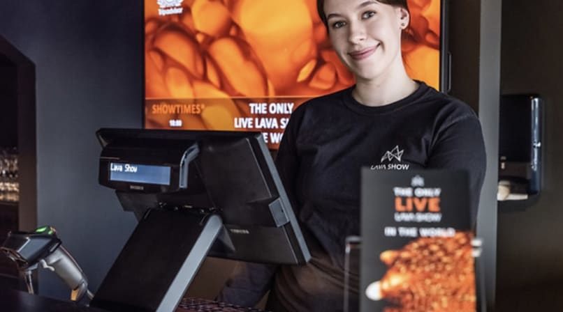 A smiling staff member in a Lava Show t-shirt stands at the ticket and merchandise counter, ready to welcome guests.