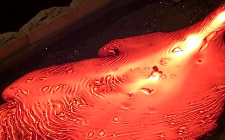 A close-up image of molten lava flowing across a dark sandbed, showing glowing orange textures, swirling lines, and bubbles as the lava spre