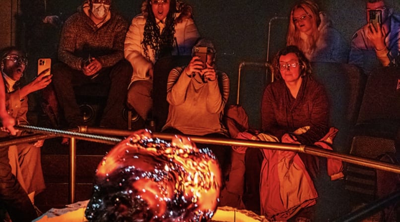 Audience members sit in close proximity watching a large, glowing, freshly-cooled lava formation, with several people photographing the mome