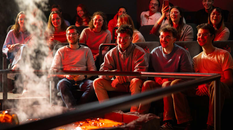 An audience seated in a dark theater watches molten lava being poured into a trench, glowing yellow-orange under stage lights.