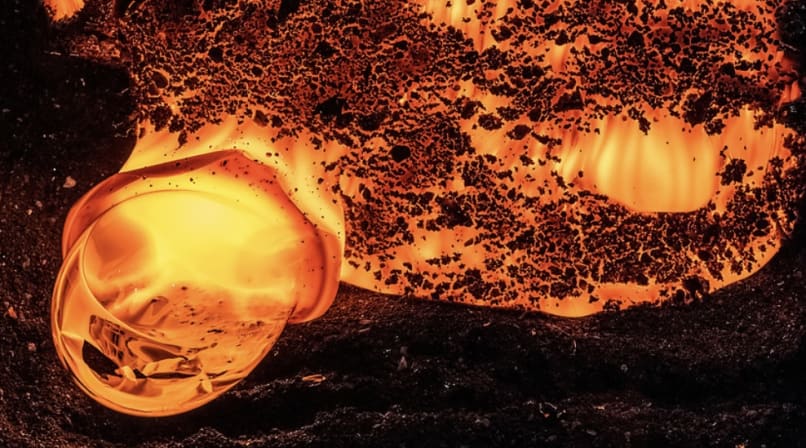 Close-up of a glowing molten lava formation bubbling on a black sandy surface, captured in vivid orange and red tones.