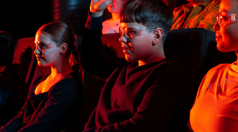 A young boy wearing safety glasses raises his hand while sitting in a dark theater alongside other children and adults, illuminated by the r
