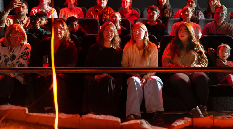 Audience members watch closely and take photos as a large, glowing blob of cooled lava rests on a sand platform in front of them.