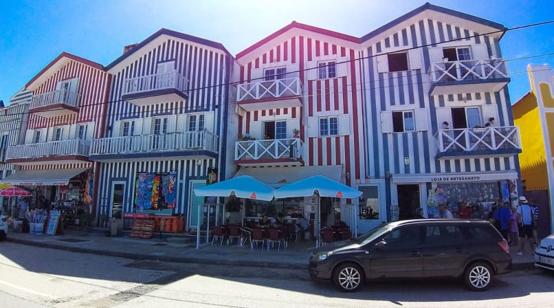 Striped houses in Costa Nova with cafés, shops, parked cars, and blue sky