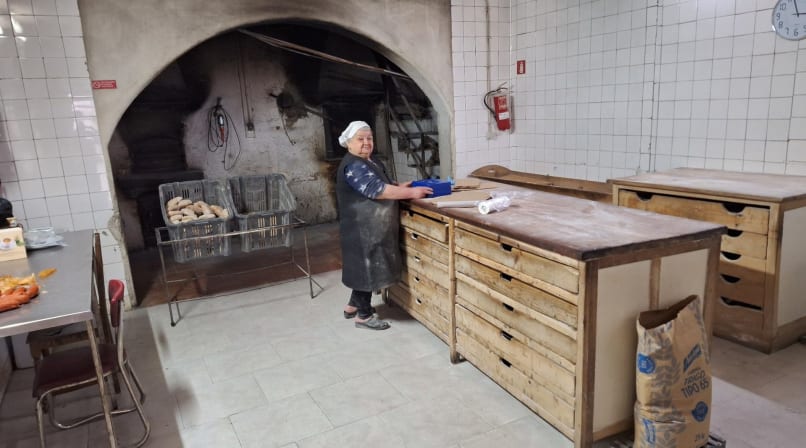 Favaios bakery kitchen with baker beside wooden worktables, bread crates, tiled walls, and traditional oven