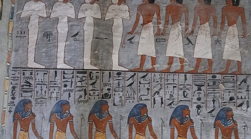 Two tiers of Ancient Egyptian figures and deities painted in blue, red, and white on a stone wall
