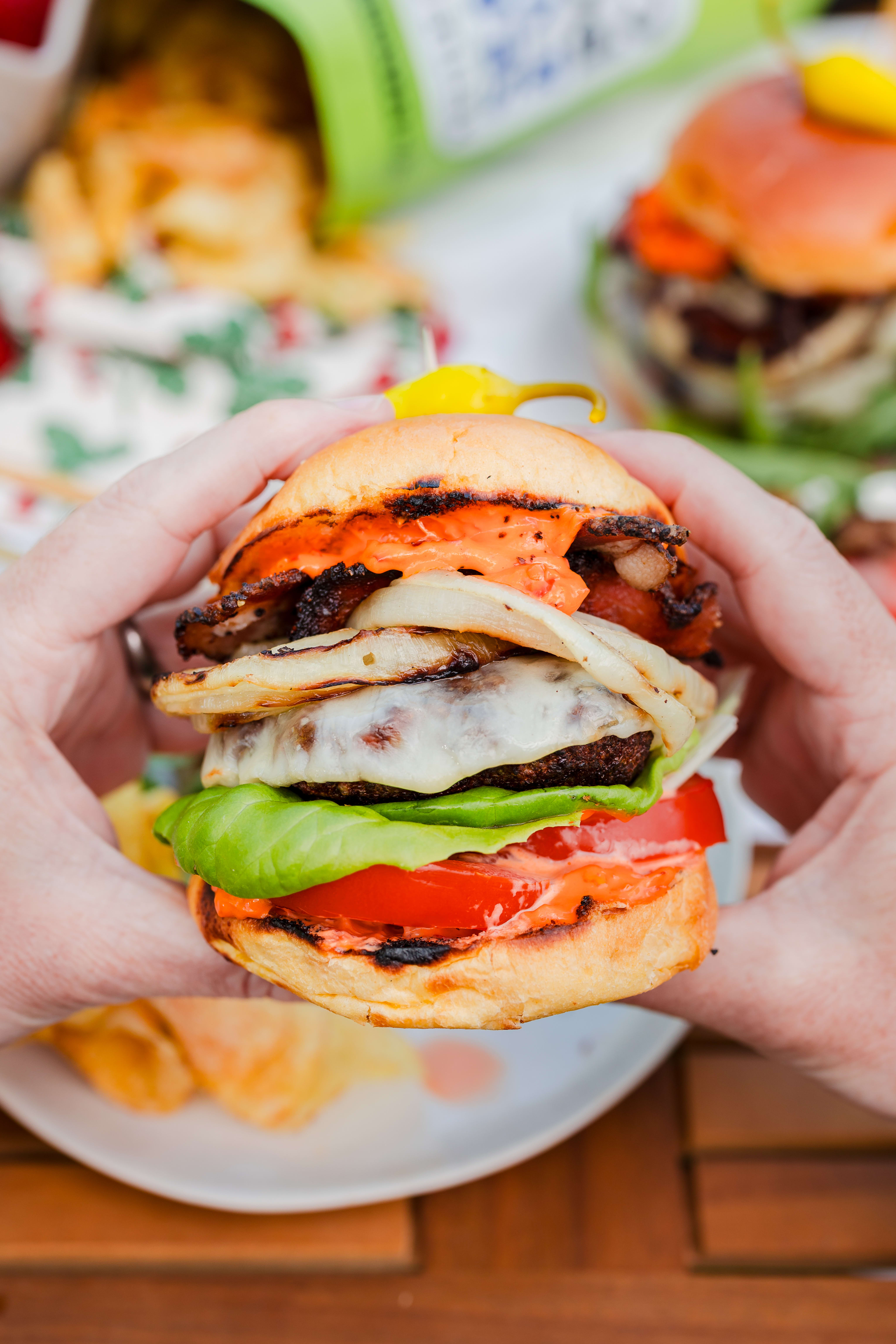 Two hands holding a burger with cheese, tomatoes, grilled onions, lettuce and Calabrian Chili mayo