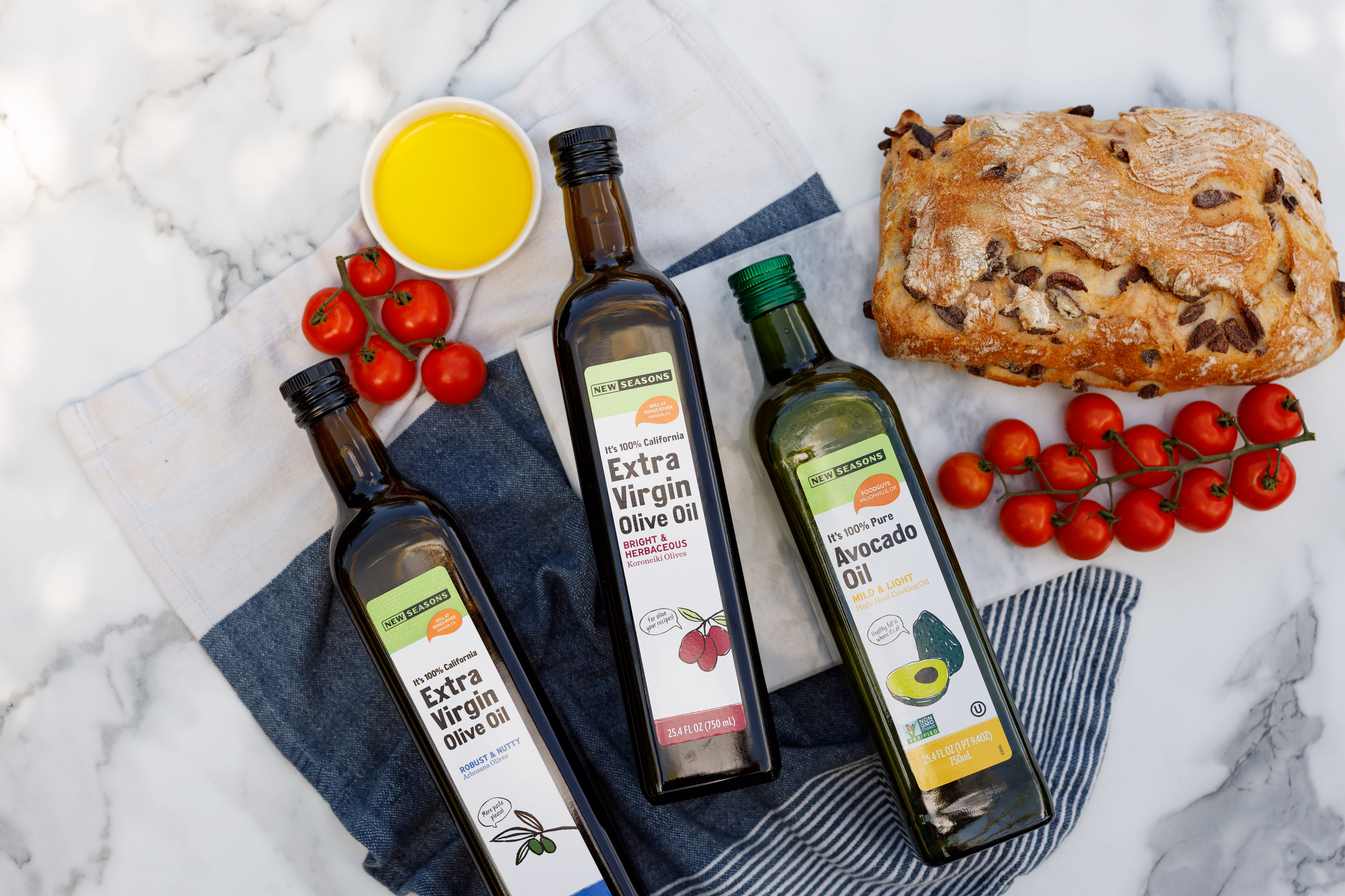 Image of New Seasons Extra Virgin Olive OIl and Avocado Oil styled with tomatoes and bread on a marble counter.