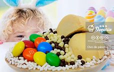 Uma criança com orelha de coelhinho apoiada sobre uma mesa observa um ovo de Páscoa de colher feito feito de chocolate branco, coberto com pedaços de bombom Ouro Branco, bolinhas de Choco Ball, granulado branco e M&M's