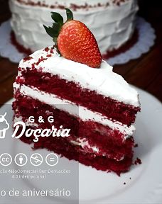 Fatia de bolo Red Velvet com cobertura branca de Cream Chesse e um morango enfeitando o topo da fatia. Fatia esta sobre um prato branco posicionado em frente ao restante do bolo.