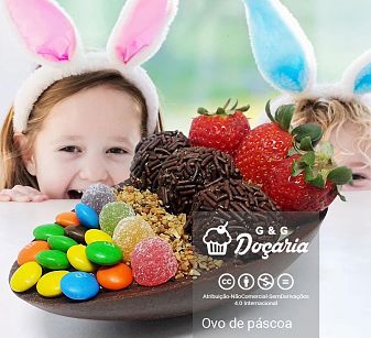 Duas crianças com orelhas de coelhinho apoiadas sobre uma mesa observam ansiosas um ovo de Páscoa de colher feito de chocolate com morangos, brigadeiros, castanhas trituradas, balas de goma e M&M's, que está sobre a mesa