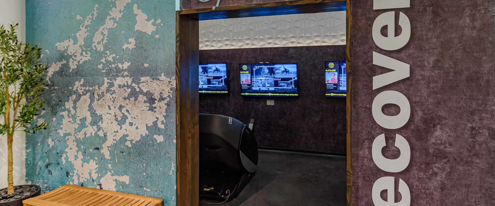 Recovery area at Gold’s Gym Long Beach, designed for post-workout relaxation. A rustic, textured wall with "Spa" and "Recovery" signage marks the entrance. Inside, high-tech massage chairs provide muscle relief, positioned in front of mounted screens displaying wellness content. A wooden bench and potted plant add to the calming ambiance, creating a space dedicated to rest and recovery.