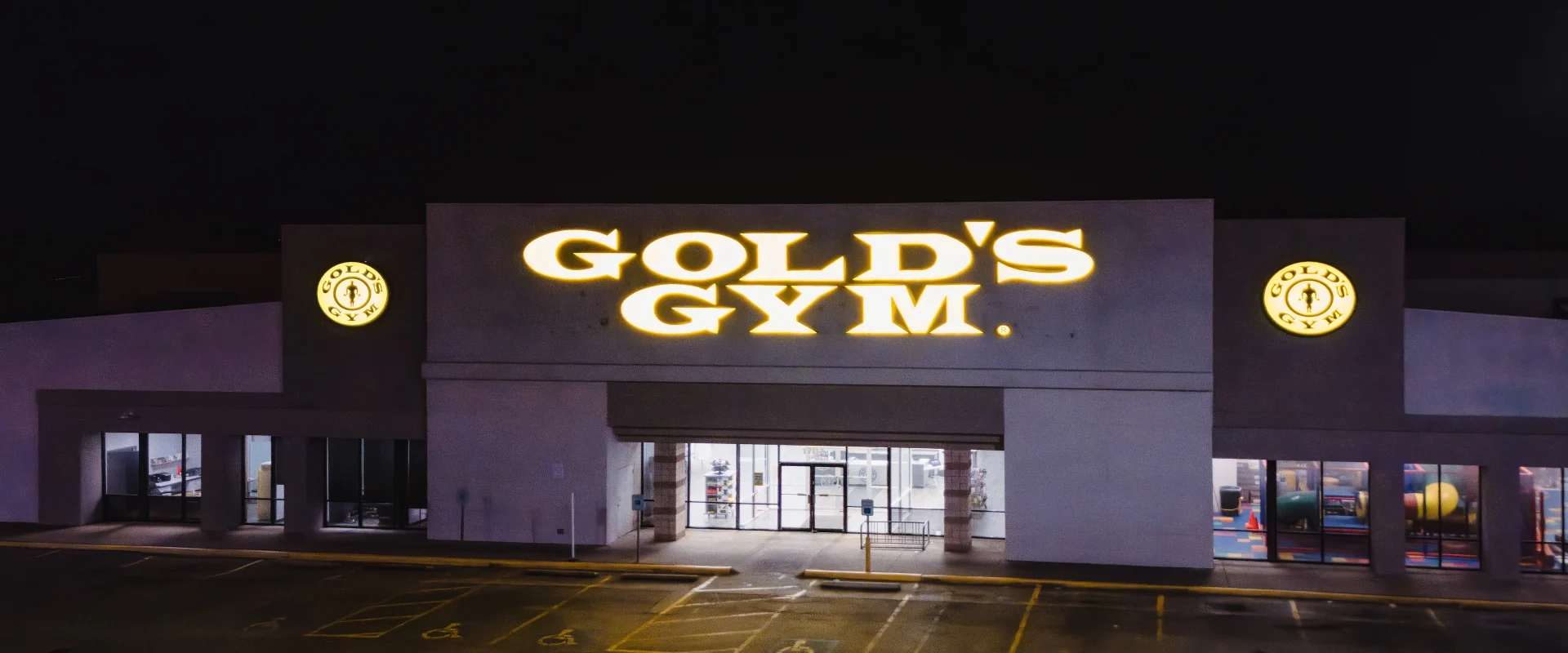 Exterior of Gold’s Gym in Austin (South Central), featuring a large illuminated sign with the Gold’s Gym logo prominently displayed above the entrance. The building is flanked by additional logo signs on both sides of the main center logo "Gold's Gym", with well-lit windows revealing interior gym areas. The parking lot includes designated handicap spaces, and the scene is set against a dark night sky.