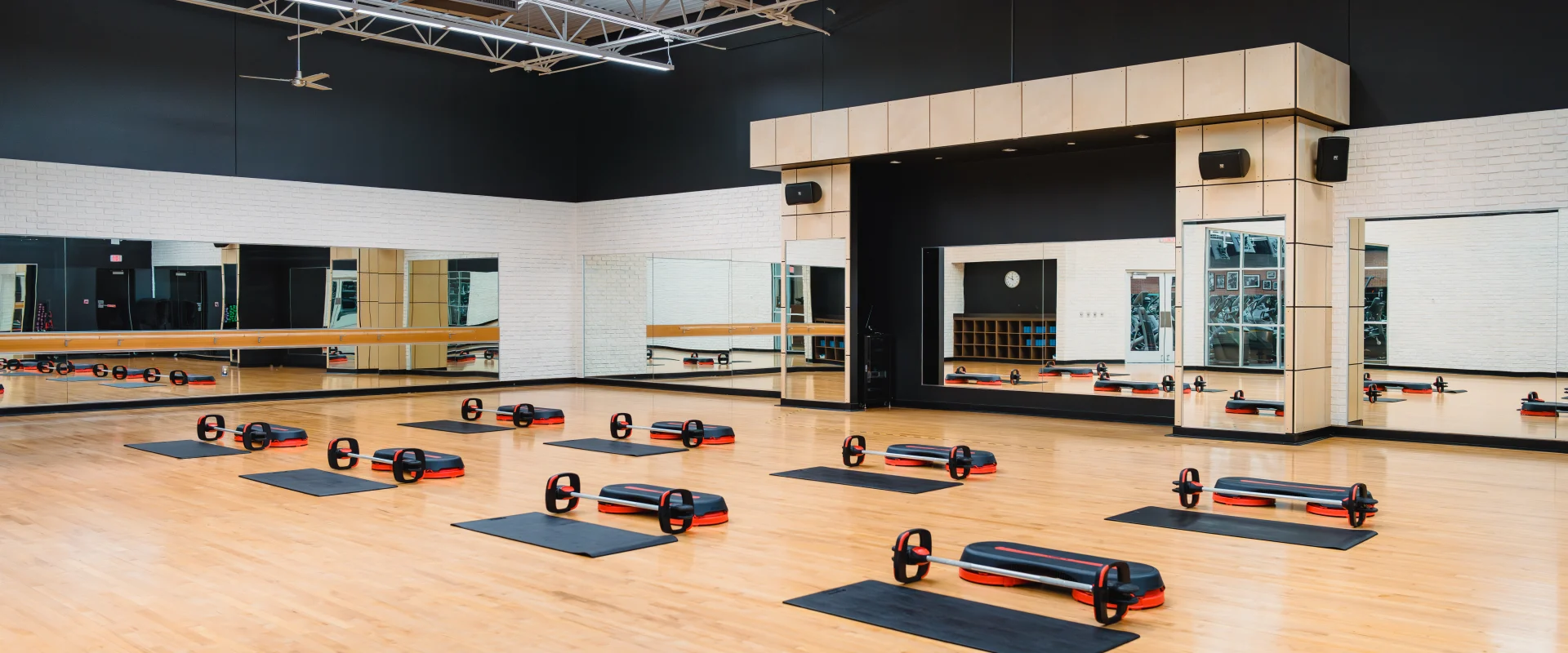 A spacious group exercise studio at Gold's Gym with wooden floors, mirrored walls, and neatly arranged steps, barbells, and mats for fitness classes.