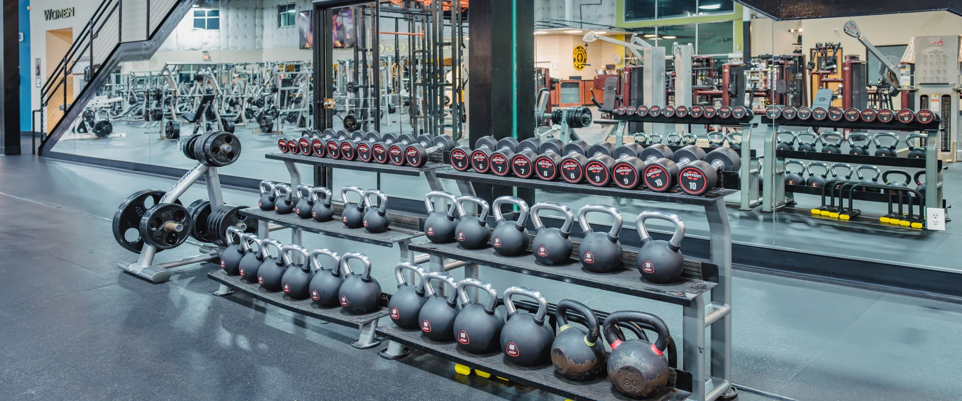 A functional training area at Gold’s Gym in Austin (South) features neatly organized kettlebells and dumbbells on silver racks against a mirrored wall. The kettlebells vary in size and weight, with some showing signs of use, while the dumbbells are uniformly branded with red and black labels. To the left, a vertical rack holds weight plates. In the background, a staircase with a black railing leads to an upper level, and gym equipment fills the open space. Bright overhead lighting reflects off the polished flooring, creating a clean, modern workout environment.