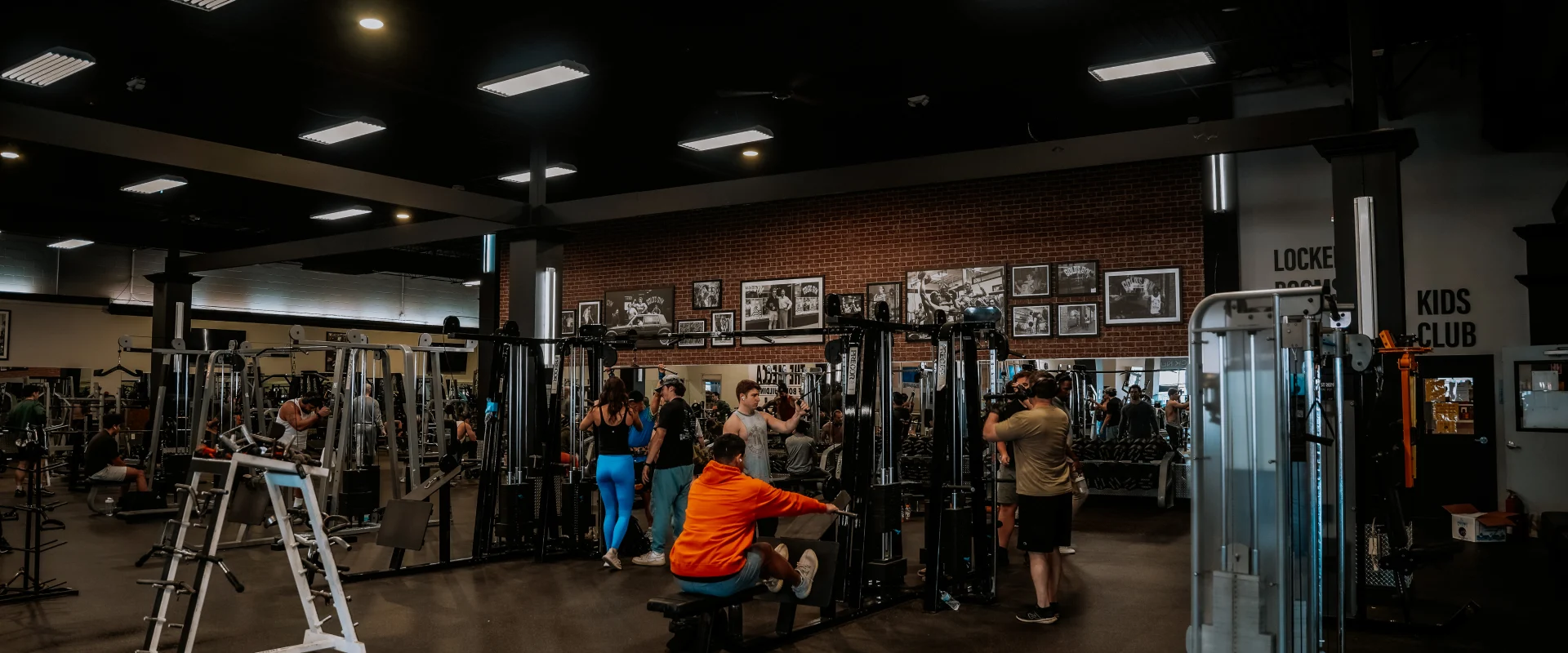 Strength training area at Gold's Gym Murfreesboro, Tennessee, featuring cable machines, free weights, and various strength equipment. Gym-goers are actively working out in a room with brick accent walls adorned with framed fitness photos. Overhead lighting illuminates the space.
