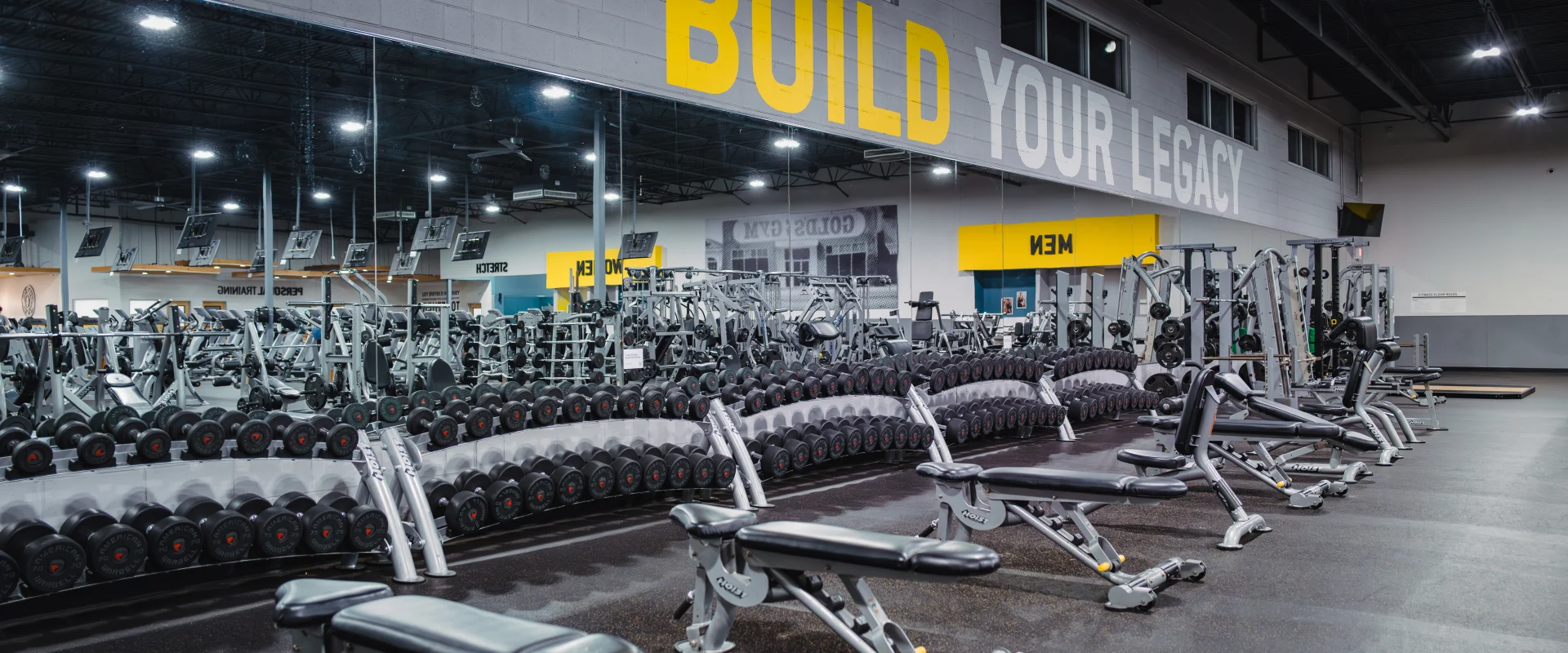 Dumbbell area at Gold's Gym Belterra featuring rows of dumbbells organized on sleek racks in front of a mirrored wall. Adjustable benches are spaced for individual workouts, with the motivational phrase "Build Your Legacy" prominently displayed on the wall above. The area is well-lit, creating a dynamic and empowering atmosphere.
