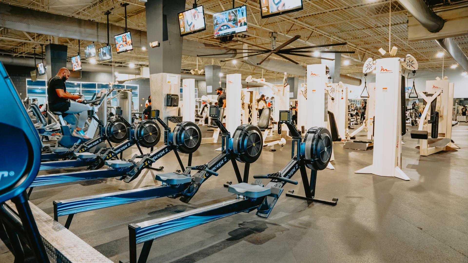 Rowing machine area at Gold’s Gym Tennessee, Lee Highway, showcasing a row of sleek rowing machines placed on a polished rubber floor. Overhead TVs display workout programs, while a high ceiling with exposed beams and ducts enhances the spacious feel. Surrounding the area are various other fitness equipment and machines, with gym members actively using the space. Bright lighting ensures the area is well-illuminated for an energetic workout environment.