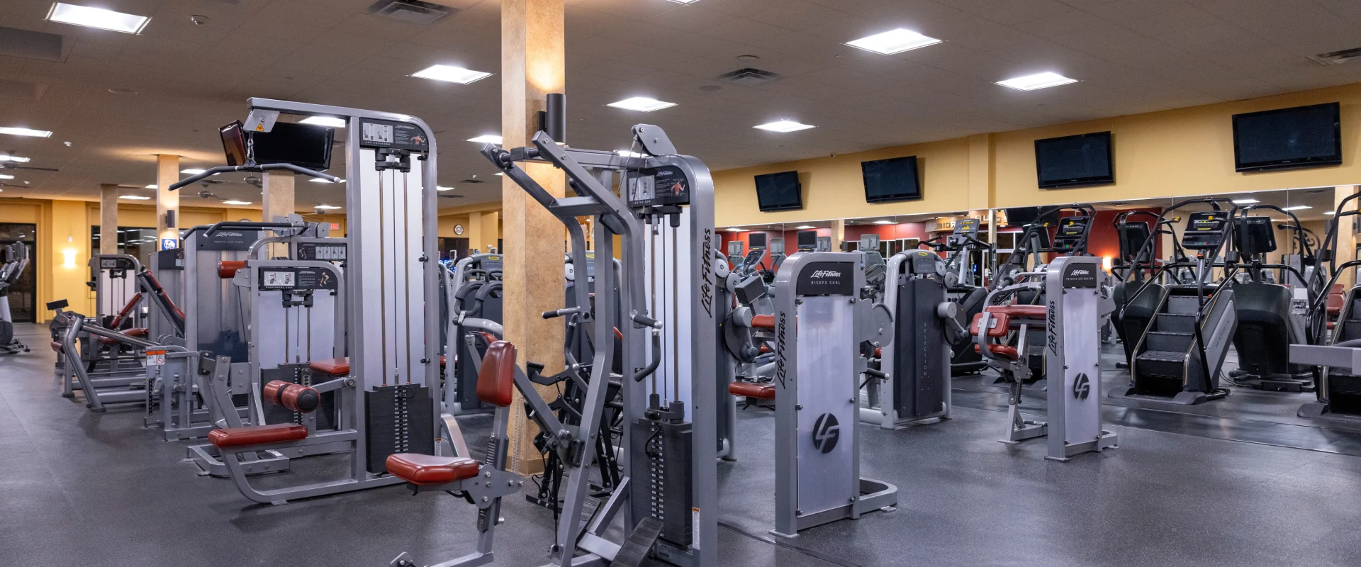 Strength equipment area at Gold’s Gym in East Northport, New York, featuring Life Fitness selectorized machines for targeted muscle training, including leg extension, bicep curl, and chest press machines. The space is organized with rubber flooring, mirrored walls, and overhead TVs lining the perimeter. Bright ceiling lights enhance visibility, while Matrix stair climbers are visible in the background for cardio integration.